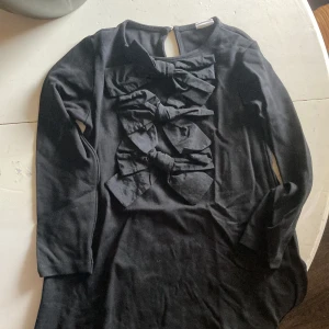 Svart klänning med stora rosetter Zara - Svart klänning från Zara med lång ärm och tre stora, snygga rosetter framtill. Rund halsringning och knapp i nacken. Mjuk och bekväm bomullskvalitet, perfekt för dig som gillar statement-detaljer och vill sticka ut lite extra.