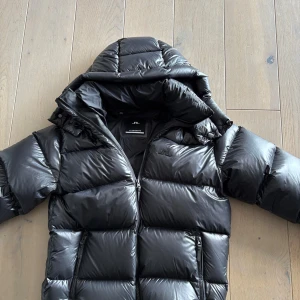 Svart pufferjacka från J.Lindeberg - Säljer en svart pufferjacka från J.Lindeberg med glansig finish och huva. Jackan har dragkedja framtill, två fickor med dragkedja och är riktigt varm och skön för kalla dagar. Typ aldrig använd, köptes i vintras. Storlek S