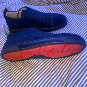 Mörkblå mocka sneakers från Louboutin - Säljer ett par mörkblå sneakers från Christian Louboutin i lyxig mocka med klassisk röd sula och snörning. Skorna har rund tå och diskret broderad logga på hälen. Insidan är i beige skinn för extra komfort. Perfekta för dig som vill sticka ut med exklusiv stil.