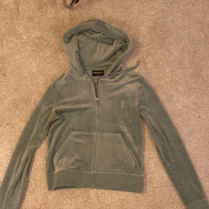 Grön velour hoodie från Juicy Couture - Säljer en ikonisk ljusgrön hoodie från Juicy Couture i mjuk velour. Tröjan har dragkedja med J-detalj, huva och känguruficka framtill. Broderad JC-logga på bröstet och ribbade muddar. Perfekt för en chill och trendig look.