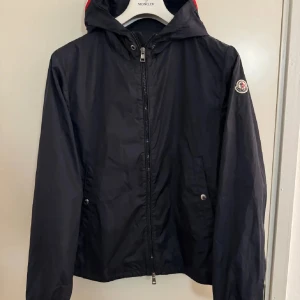 Svart vindjacka från Moncler med huva - Navy vindjacka från Moncler med klassisk logga på ärmen och huva med rödvit detalj. Jackan har dragkedja framtill, två fickor med tryckknappar och en stilren, sportig look. Perfekt för dig som gillar exklusiva streetwear-vibbar. perfekt till hösten