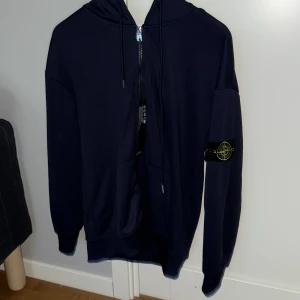 Mörkblå hoodie från Stone Island - Säljer en mörkblå hoodie från Stone Island med dragkedja, huva och klassisk logga på ärmen. Helt oanvänd, skriv vid frågor eller funderingar, pris kan diskuteras