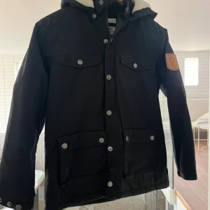 Svart parkajacka från Fjällräven - Svart parkajacka från Fjällräven med huva och mysigt vitt teddyfoder. Jackan har flera fickor med tryckknappar och Fjällräven-logga på ärmen. Perfekt för kalla dagar och outdoor-häng.