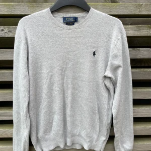 Grå tröja från Polo Ralph Lauren - Snygge ljusgrå tröja från Polo Ralph Lauren i mjuk pima bomull. Klassisk rund halsringning, långärmad och med den ikoniska broderade loggan på bröstet. Perfekt för dig som gillar stilren och enkel design. Prima Cotton är det motsvariga för lyxigare och bättre material, känns som merinoull! Axelbredd 45,5 cm och längd på tröjan är 61 cm 