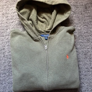 Olivgrön hoodie från Polo Ralph Lauren - Olivgrön hoodie från Polo Ralph Lauren med dragkedja och huva. Klassisk orange broderad logga på bröstet och känguruficka framtill. Tillverkad i mjuk bomull.