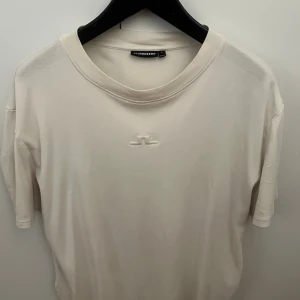 Vit t-shirt från J.Lindeberg - Snygg vit t-shirt från J.Lindeberg med diskret broderad logga på bröstet. Klassisk rund halsringning och korta ärmar. Tillverkad i mjuk bomull för en skön känsla. Perfekt för en clean och stilren look.