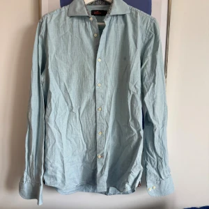 Ljusblå skjorta från Morris - Snygg ljusblå skjorta från Morris med klassisk krage och diskret broderad logga på bröstet. Skjortan har långa ärmar, knappar framtill och är tillverkad i mjuk bomull. Perfekt för dig som gillar stilrena och enkla plagg.