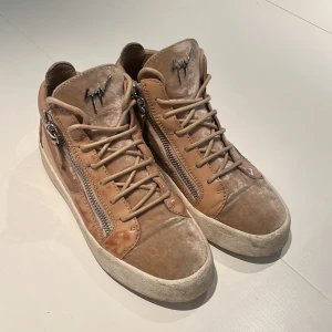 Beige sneakers från Giuseppe Zanotti - Säljer ett par snygga beige sneakers från Giuseppe Zanotti med coola dragkedjor på sidorna, snörning och logga i metall framtill. Skorna har en mix av sammet och lackdetaljer som ger en lyxig vibe. Perfekta för dig som vill sticka ut med din stil. Är även öppen för byten 