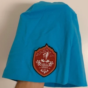 Turkos mössa från Muddus med Fjällgården-patch - Turkos mössa från Muddus med en stor röd Fjällgården Åre-patch framtill. Tillverkad i mjuk bomull med lite stretch för skön passform. Perfekt för dig som vill sticka ut med färg och cool badge på vintern eller våren.