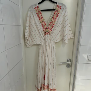 Vit långklänning med broderi och guldtrådar - Lång, vit klänning i boho-stil med guldiga ränder, v-ringning och broderade blommor i rosa, rött och beige längs halsringningen. Vida ärmar, knytband med tofsar i midjan och volangkant nertill. 