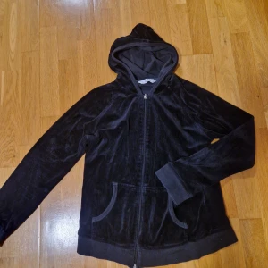 Svart velour hoodie från Zona - Mysig svart hoodie från Zona i mjuk velour med dragkedja framtill och två fickor. Dragkedjan saknar sin yttersta del som syns på bilden men fungerar bra endå. Tröjan har huva och långa ärmar med ribbade muddar.