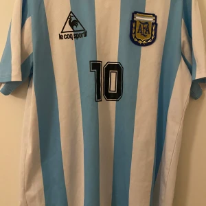 Retro Maradona 1986 VM Argentina tröja - Klassisk Argentina fotbollströja från VM 1986 med ljusblå och vita ränder, nummer 10 och Maradona på ryggen. Broderad AFA-logga på bröstet och Le Coq Sportif-märke. Kortärmad modell i lätt och ventilerande material, perfekt för retro fotbollsfans.