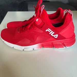 Röda sneakers från Fila - Snygga röda sneakers från Fila, Storlek 43,  med vit logga på sidan och tungan. Ovandelen är i mesh och syntet för bra andningsförmåga och komfort. Skorna har snörning och en vit och röd sula med sportig look. Perfekta för dig som vill sticka ut med färg på fötterna.