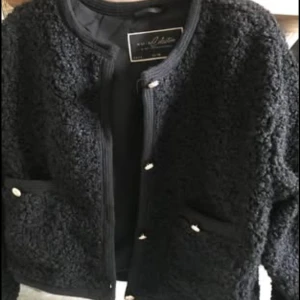 Svart teddyjacka med guldknappar - Säljer en svart teddyjacka från Moliin med rund halsringning och snygga guldknappar framtill. Jackan har två fickor framtill och är tillverkad i ett mjukt, fluffigt material som ger en cool och trendig look. Perfekt för dig som gillar stilrena och mysiga plagg.