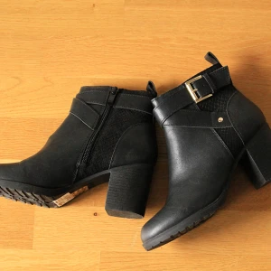 Svarta klackskor - Snygga svarta skor med chunky klack, rund tå och guldspänne. Skorna är i skinnimitation och har dragkedja på insidan och mönstrade partier. Perfekta för en snygg höstlook! Enbart provade, aldrig använda. 