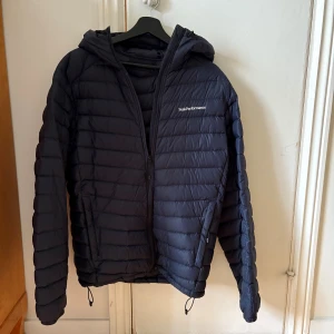 Dunjacka Peak Performance  - Tunnare blå dunjacka med huva, två sidofickor med dragkedja samt innerficka med dragkedja. 