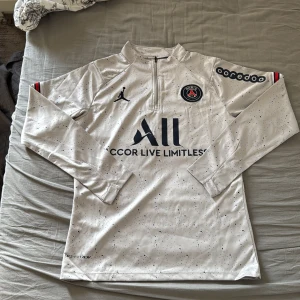 PSG långärmad träningströja Jordan S - Snygg långärmad PSG träningströja från Jordan i vit med svarta prickar. Tröjan har halv dragkedja, klubbmärke på bröstet och tryck på ärmen. Tillverkad i lätt och ventilerande Dri-FIT material, perfekt för fotbollsträning.