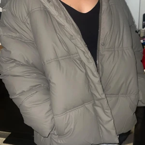 Grå pufferjacka med huva - Säljer en grå pufferjacka med huva och dragkedja. Jackan är quiltad och har ribbade muddar vid ärmsluten för extra komfort. Perfekt för kalla dagar och har en oversized passform som ger en chill vibe. En helt ny vinterjacka från FB Sister, köpt på NewYorker. Storlek L. Aldrig använd, endast provad hemma. Väldigt varm och bekväm, perfekt för vintern! Nypris var 600kr, säljer för 500kr. Kan skickas eller hämtas i Stockholm.