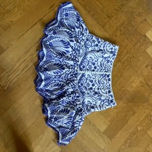 Vit och blå broderad kortkjol - Superfin kortkjol i vitt med blått broderat blommönster och volangkant. Kjolen har dekorativa knappar framtill och dragkedja bak. Materialet känns som bomull och ger en luftig känsla, perfekt för varma dagar. Snygg och unik design med detaljerade broderier. Smal i storlek