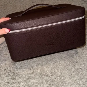 Brun necessärväska från CAIA - Stilren och rymlig brun necessärväska från CAIA i rektangulär form. Väskan har ett handtag upptill, silverfärgad dragkedja och är tillverkad i ett skinnliknande material. Insidan har fack för penslar och gott om plats för smink eller andra prylar.