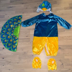 Påfågelkostym i blått och gult pelican dräkt - Färgglad påfågelkostym med glansigt blått överdel, gula byxben och avtagbar stjärt med påfågelfjädermönster. Hatten har blå fjädrar och grönt hjärta. Kostymen är i mjuk polyester och har tryckknappar för enkel påklädning. Perfekt för maskerad eller temadagar / halloween 🎃 🥰inköpspris: 599:- 