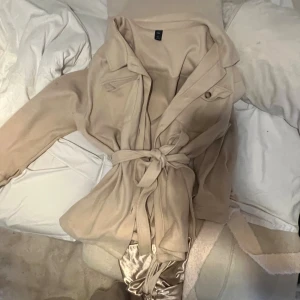 Beige och svart kappa med bälte för 100kr - Två stilrena kappor i beige och svart med klassisk krage och knytskärp i midjan. Båda har knappar framtill och en rak, längre passform som ger en clean look. Perfekt för dig som gillar minimalistisk stil och vill ha en jacka som funkar till många outfits.