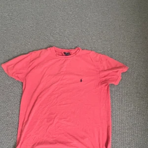 Rosa t-shirt från Polo Ralph Lauren - Säljer en klassisk röd t-shirt från Polo Ralph Lauren med broderad marinblå logga på bröstet. T-shirten är i mjuk bomull och har en avslappnad passform med korta ärmar och rund hals. Perfekt för en chill och stilren look.