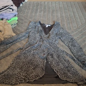 Leopardmönstrad omlottklänning NLY Trend - Snygg kort omlottklänning från NLY Trend i storlek 36. Klänningen har leopardmönster i svart och beige, långa ärmar och ett knytband i midjan. Materialet är lätt och luftigt, perfekt för att sticka ut med en cool v-ringad look.