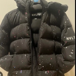 Svart pufferjacka från NVLTY med färgstänk - Säljer en svart pufferjacka från NVLTY med coola färgstänk i vitt, rosa och blått. Jackan har huva, dragkedja och en oversized vibe. Perfekt för dig som vill sticka ut i vinterkylan.