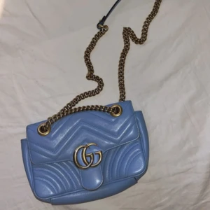 Blå Gucci Marmont axelväska i skinn - Supersnygg ljusblå Gucci Marmont axelväska i skinn med guldfärgad kedja och GG-logo framtill. Väskan har quiltad design med hjärtdetalj på baksidan och klassisk flap. Skriv för fler bilder, köpt för 4 år sen så kvitto och box finns inte där av priset men QR/kod finns, finns små defekter skickar bild 