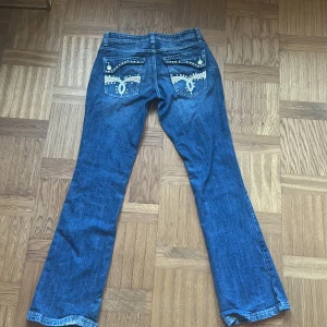Blå bootcut jeans från Bershka strl 34 - Snygga blå jeans från Bershka i bootcut-modell med coola detaljer på bakfickorna, som broderier och nitar. Jeansen har en glittrig knapp framtill och klassisk femficksdesign. 