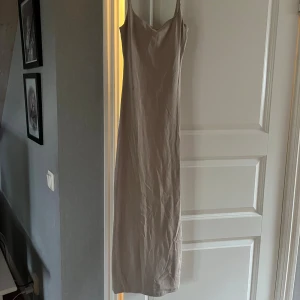 Beige långklänning med smala axelband - En stilren beige långklänning med smala axelband och rak passform. Klänningen har en enkel design utan mönster och är tillverkad i ett mjukt material som känns skönt mot huden. Perfekt för dig som gillar minimalistisk stil.