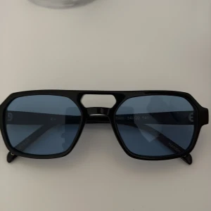Svarta solglasögon Corlin Eyewear - Snygga svarta solglasögon från Corlin Eyewear med blåtonade glas och en modern, kantig form. Bågarna är i blank svart plast och har en cool detalj med öppning vid näsryggen. Perfekta för dig som vill sticka ut med en trendig accessoar.