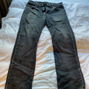 Levi's 501 svarta jeans W30 L30 - Klassiska svarta Levi's 501 jeans med raka ben och knappgylf. Jeansen har fem fickor och den ikoniska läderpatchen bak i midjan. Perfekt för dig som gillar en tidlös och avslappnad stil. Materialet är robust denim i bomull. Litet hål i skrevet men enkelt att lösa. 