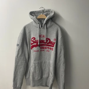 Grå Superdry hoodie med röd logga - Grå hoodie från Superdry med stor röd broderad logga på bröstet och japanska tecken. Klassisk känguruficka, snörning i huvan och ribbade muddar. Perfekt för dig som gillar streetwear och vill ha en chill look.