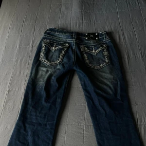 Miss Me blå jeans med broderade fickor - Snygga blå jeans från Miss Me med slitningar och kontrastsömmar. Bakfickorna har detaljerade broderier och nitar, samt en metallogo. Jeansen har bootcut-modell och är tillverkade i klassiskt jeanstyg.