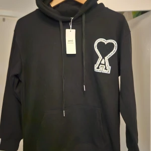 Svart hoodie från Ami Paris - Svart hoodie från AMI Paris, Alexandre Mattiussi, med A-loggan på bröstet. Hoodien har en justerbar huva med dragsko och en känguruficka fram. Ej använd.  Storlek small. Mått taget med hoodien hängande på galgen så därför ej exakt på mm.  Axeln ner till muddslut: 57 cm Rakt över (utsträckt), armhåla till armhåla: 50 cm Längd, nacke (söm) till längst ner: 61 cm 
