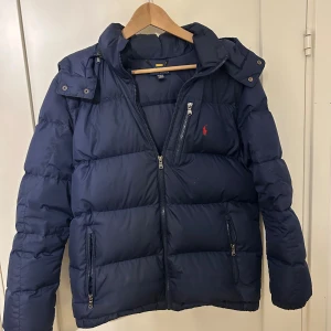 Ralph lauren jacka - Säljer en mörkblå pufferjacka från Polo Ralph Lauren med huva och dragkedjor framtill. Jackan har det klassiska röda Polo-loggan broderad på bröstet och flera praktiska fickor med dragkedja. Perfekt för kalla dagar och riktigt snygg streetstil.