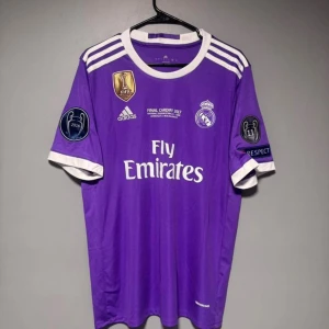Lila Real Madrid fotbollströja Adidas - Snygg lila Real Madrid fotbollströja från Adidas med vita detaljer och tryck. Tröjan har klubbmärke, sponsorlogga och Champions League-patchar på ärmarna. Perfekt för dig som vill sticka ut på planen eller visa ditt stöd för laget.