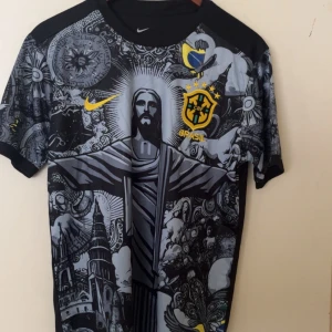 Svart fotbollströja - Unik svart fotbollströja med grått och gult tryck. Framsidan har detaljerad grafik med Kristusstatyn, kyrka och konstnärliga mönster. Gult märke och logga på bröstet. Korta ärmar med tryck, baksidan är enkel med emblem.