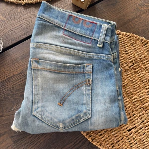Ljusblå slitna jeans från Dondup - Säljer ett par blå jeans från Dondup i storlek 30. Jeansen har slitna detaljer och hål på benen för en cool, avslappnad look. Klassisk femficksmodell med knapp. Perfekta för dig som gillar streetstyle och en edgy vibe. Obs defekt i skrevet 