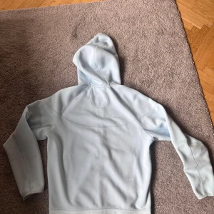 Ljusblå Nike NOCTA hoodie med dragkedja - Säljer en ljusblå hoodie från Nike NOCTA med hel dragkedja framtill och två fickor med dragkedja. Tröjan har huva, långa ärmar och diskreta loggor på bröstet och ärmen. Perfekt för dig som gillar streetwear och sportig stil.