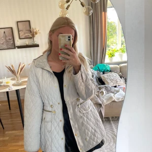 Quiltad beige jacka med huva, M - Säljer en quiltad beige jacka med huva och guldfärgade detaljer. Jackan har dragsko i midjan, fickor med både dragkedja och knappar samt långa ärmar. Perfekt för dig som vill ha en snygg och praktisk jacka till höst och vår. Storlek M . Köpt vintage så vet inte märke 