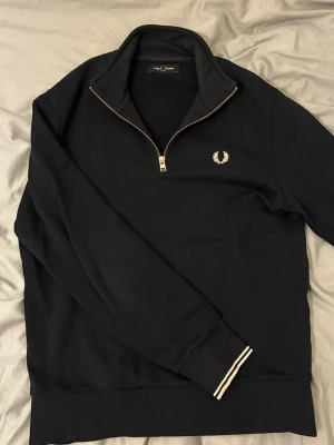 Svart half zip tröja från Fred Perry - Snygg svart tröja från Fred Perry med half zip och klassisk logga broderad på bröstet. Tröjan har långärmade ribbade muddar med vita ränder och en hög krage. Perfekt för dig som gillar stilrena och sportiga plagg.