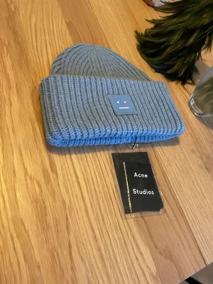 Ljusblå pansy beanie från Acne Studios - Säljer en snygg blå stickad mössa från Acne Studios med det klassiska ansikts-emblemet framtill. Mössan har bred uppvikt kant och är gjord i mjukt material som håller dig varm. Perfekt för att addera lite stil till din outfit.