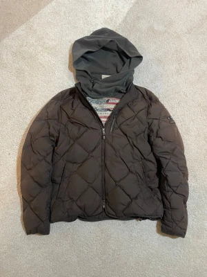 Mörkbrun quiltad pufferjacka Morris | S - Quiltad pufferjacka från Morris i mörkbrunt med snygg huva och dragkedja framtill. Jackan har ett rutmönstrat yttertyg och är fodrad för extra värme. Nypris cirka 2200 
