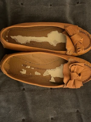 Bruna loafers med rosett - Snygga bruna loafers med rund tå och dekorativ rosett framtill. Skorna har en platt sula och är tillverkade i skinn med sömmar längs kanterna. Perfekta för dig som gillar klassisk och enkel stil med en twist.