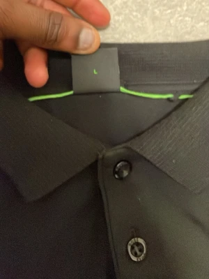 Svart polotopp från BOSS, slim fit - Svart polotopp från BOSS i slim fit med lyxig bomullskänsla. Toppens vänstra sida har en snygg detalj med BOSS-loggor i ton-i-ton. Klassisk krage och knäppning framtill, samt diskret broderad BOSS-text på bröstet. Perfekt för dig som gillar stilrena och moderna plagg.