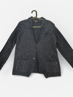 Mörkblå stickad cardigan - Snygg mörkblå stickad kavaj från Gaudì Jeans med vitmelerad struktur. Två framfickor, klassisk krage och två knappar framtill. Perfekt för dig som gillar en chill men stilren look. Passar till både jeans och chinos. Knappt använd och perfekt som julklapp. Fraktar samma dag som köp!