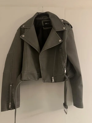Grå skinnjacka från FB Sister - Säljer en grå skinnjacka från FB Sister med klassisk bikerstil. Jackan har silvriga dragkedjor, bred krage med tryckknappar och ett bälte i midjan. Långärmad modell med detaljerade sömmar och fickor med dragkedja. Perfekt för dig som vill ha en edgy look.